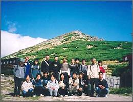 青空と山を背景に集合写真を撮る現地の学生と中学生の写真