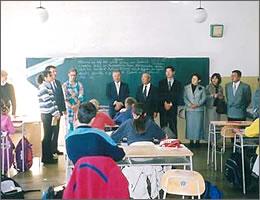 子供たちを前に黒板の前で教師の話を聞く訪問団の写真
