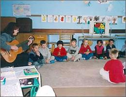 教師がギターを弾き、子供たちがじゅうたんの上に座って歌を歌っている写真