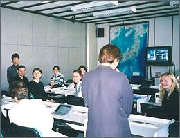 モニターに日本地図が映された教室で現地の学生に混じって自己紹介する訪問団の写真